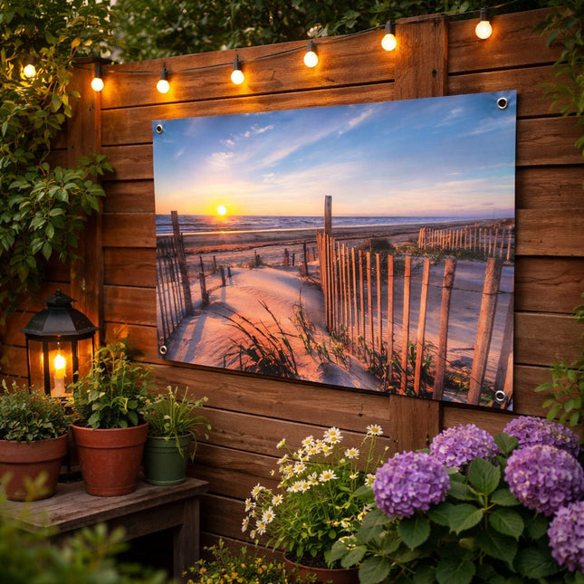 Tuinposter met duinstrand en zonsondergang bevestigd aan houten schutting in een sfeervolle tuin met planten en verlichting.